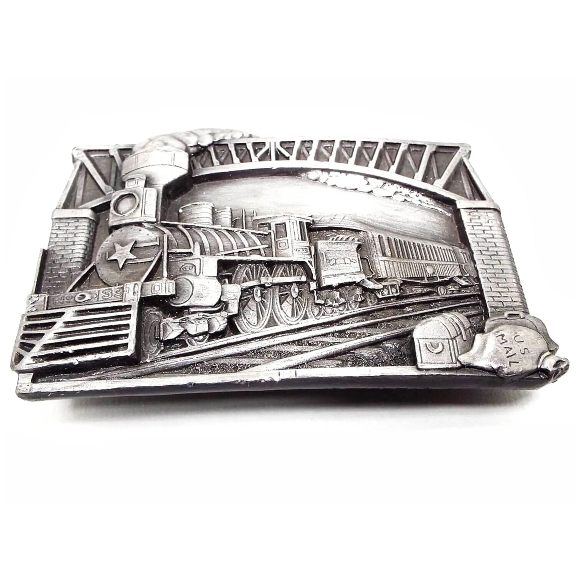 Front view of the retro vintage Bergamot Brass Works train belt buckle. The metal buckle is pewter with varying shades of gray. The raised design shows a steam engine going under a bridge. Down by the tracks on the right side of the buckle is a treasure chest, a luggage bag, and a sack laying on its side that says US Mail. The locomotive has a star on the front and looks to have No 33 or No 39 on the bottom. 