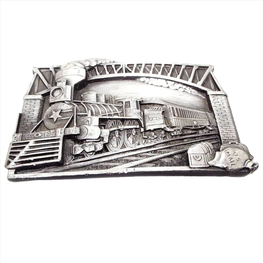 Front view of the retro vintage Bergamot Brass Works train belt buckle. The metal buckle is pewter with varying shades of gray. The raised design shows a steam engine going under a bridge. Down by the tracks on the right side of the buckle is a treasure chest, a luggage bag, and a sack laying on its side that says US Mail. The locomotive has a star on the front and looks to have No 33 or No 39 on the bottom. 