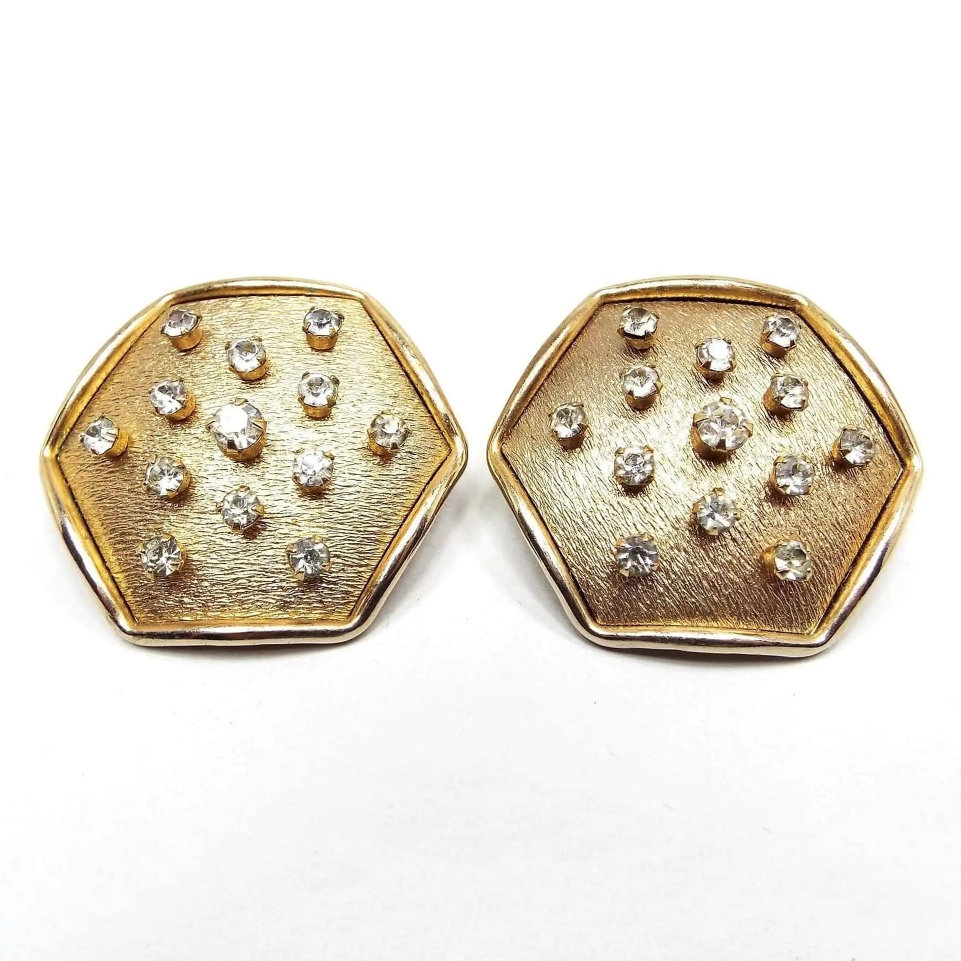 Front view of the retro vintage geometric clip on earrings. The metal is brushed matte gold tone in color with a shiny edge. They are large hexagon in shape with small round prong set rhinestones throughout the hexagon.