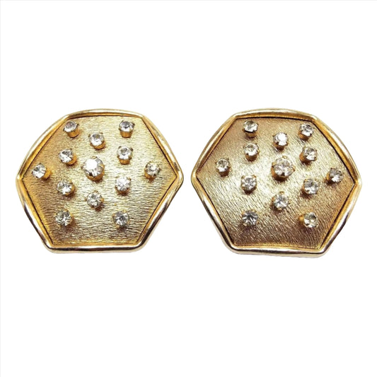 Front view of the retro vintage geometric clip on earrings. The metal is brushed matte gold tone in color with a shiny edge. They are large hexagon in shape with small round prong set rhinestones throughout the hexagon.