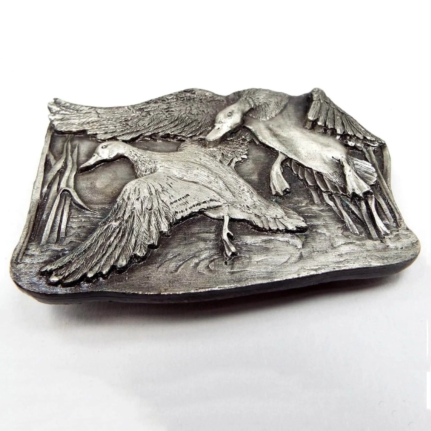 Front view of the retro vintage Bergamot Brass Works belt buckle. It depicts two flying ducks that look like they're landing on the lake. The background area has reeds and a water like design. The pewter metal of the buckle has various shades of dark and light gray and has a detailed raised 3D like design.