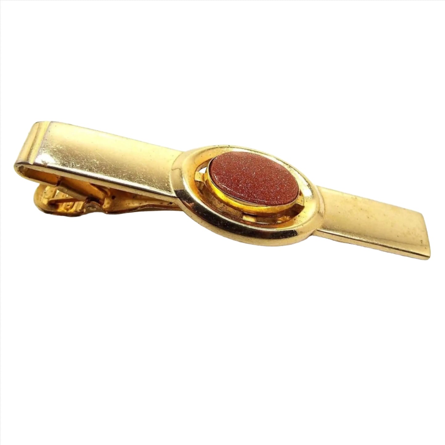 Top view of the Anson Mid Century vintage tie clip. It has a long rectangle shape with an oval in the middle. The metal is gold tone in color. The middle oval has a goldstone cab which is made of glass with tiny micro specks of copper glitter that sparkles as you move around. It is a burnt orange in color. Tie clip has an alligator style clip on the back.