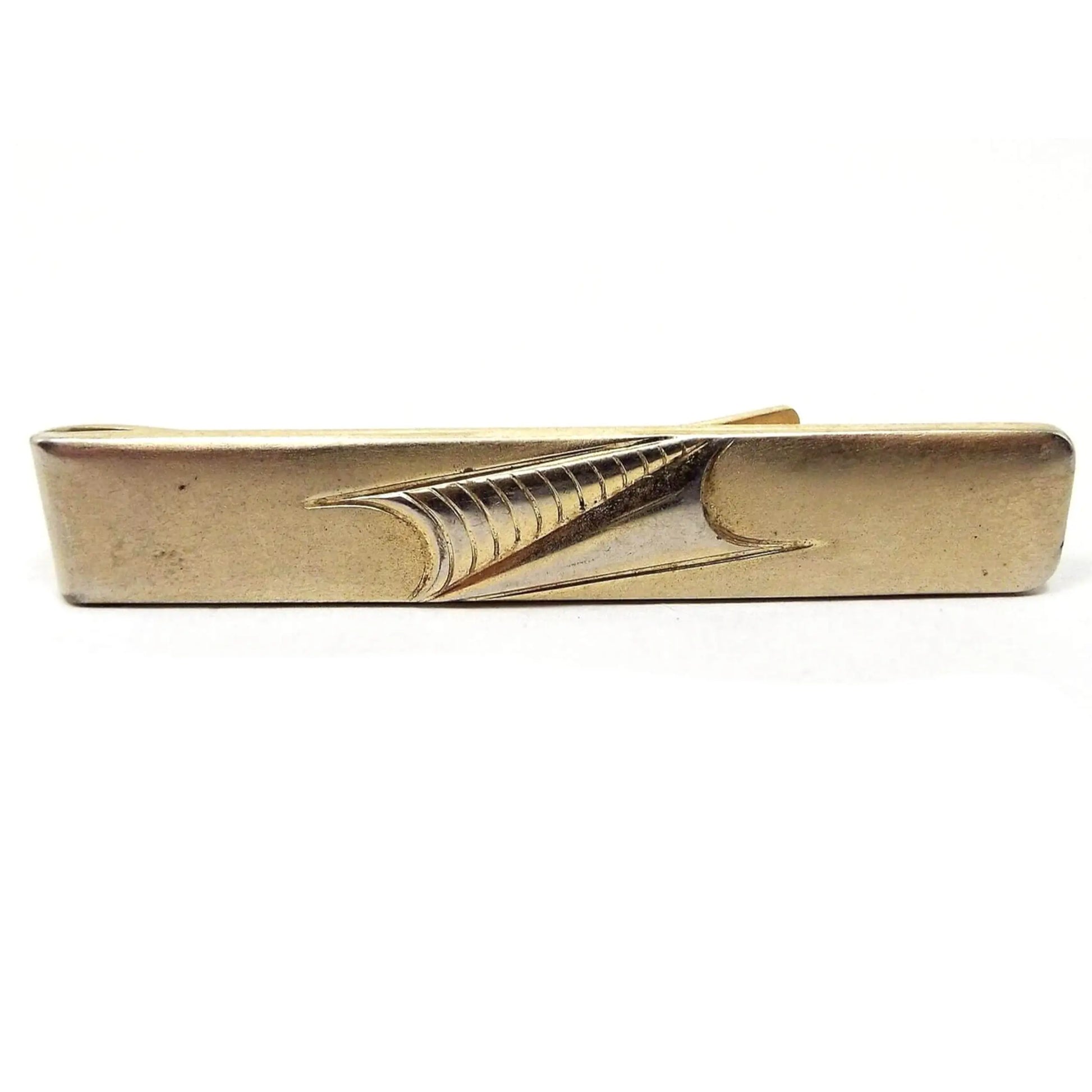Front view of the 1950's Anson vintage tie bar. Metal is darkened gold in color. It is a long rectangle shape with two modernist style triangles side by side pointing towards each other in the middle. Each triangle has curved indented bottoms. One is plain and the other has curved lines on it. 