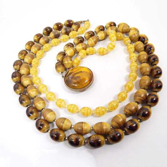 Top view of the Mid Century vintage Selini multi strand necklace. There are three strands of oval beads. The top strand has beads with a thin stripe design in shades of yellow. The middle strand has larger beads with shades of yellow and brown. The bottom strand has larger beads with shades of yellow to dark brown. There is a large flat round box clasp at the end that has a plastic domed cab with marbled shades of yellow to dark brown. The metal spacer beads and clasp setting are gold tone in color.