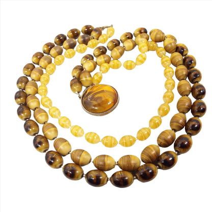 Top view of the Mid Century vintage Selini multi strand necklace. There are three strands of oval beads. The top strand has beads with a thin stripe design in shades of yellow. The middle strand has larger beads with shades of yellow and brown. The bottom strand has larger beads with shades of yellow to dark brown. There is a large flat round box clasp at the end that has a plastic domed cab with marbled shades of yellow to dark brown. The metal spacer beads and clasp setting are gold tone in color.