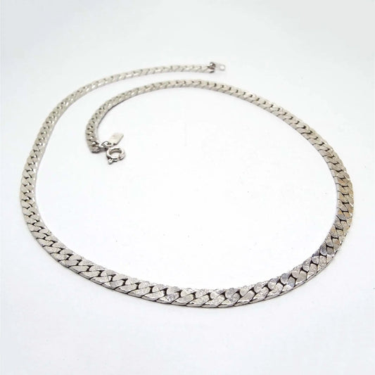 Top view of the retro vintage chain necklace. The metal is silver tone in color. The herring bone chain has lightly textured links and a round spring ring clasp at the end. There is a small rectangular hang tag by the clasp.