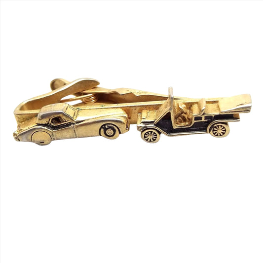 Angled front view of the retro vintage Swank car tie clip. The metal is gold tone in color. The front bar has a 3D style vintage car on the left and an antique style car on the right with a black painted body. The alligator style clip is showing on the back.