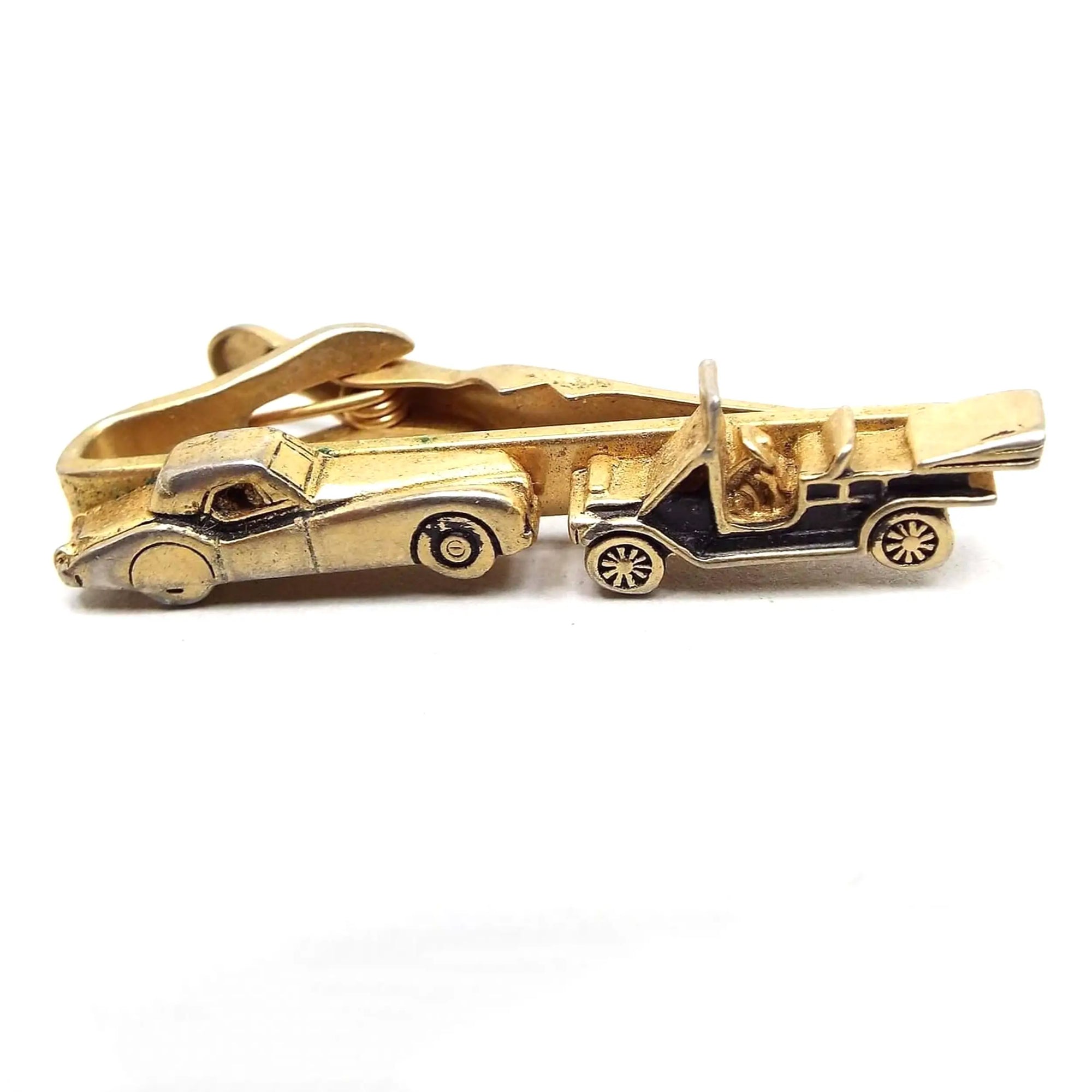 Angled front view of the retro vintage Swank car tie clip. The metal is gold tone in color. The front bar has a 3D style vintage car on the left and an antique style car on the right with a black painted body. The alligator style clip is showing on the back.