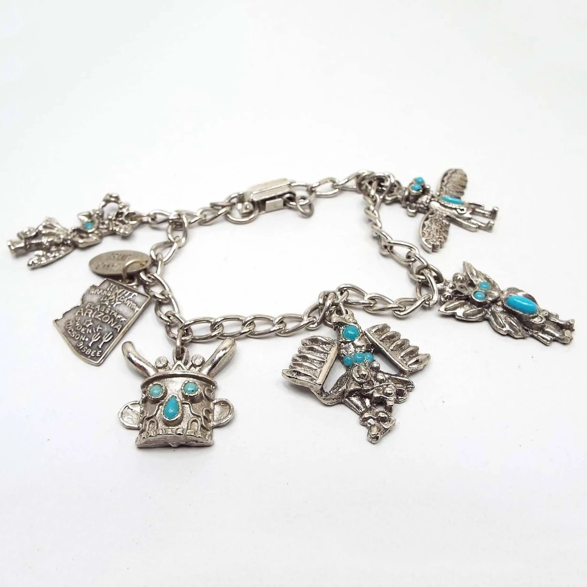 Top view of the 1970's retro vintage Arizona charm bracelet. Bracelet is silver tone in color with a curb link chain and a snap lock clasp on the end. There are 6 charms and a hang tag. The hang tag says Grand Canyon it. The charm shaped like Arizona is sterling silver. The other charms are silver plated Native American Kachina style charms with imitation turquoise cabs.