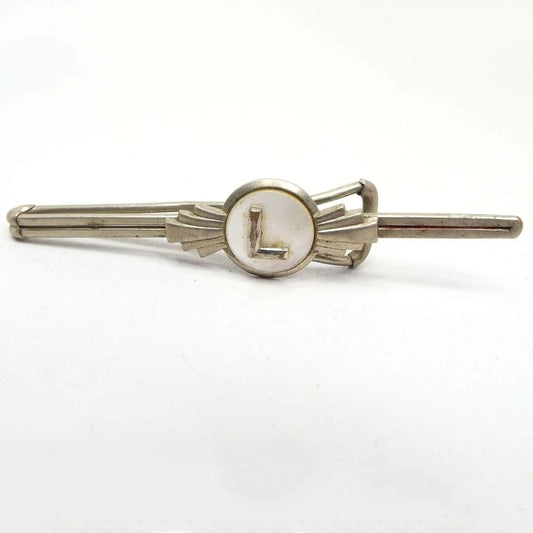 Front view of the 1930's Art Deco vintage letter tie bar. The metal is silver tone in color. The front has a long thin double wire sort of bar. In the middle of the bar is a round bezel setting with pearly white mother of pearl shell cab in the middle. There is a block letter initial L in the middle. Each side of the round area has silver tone raised flares on the sides. The back has a wider open bar for a slide on style tie bar.