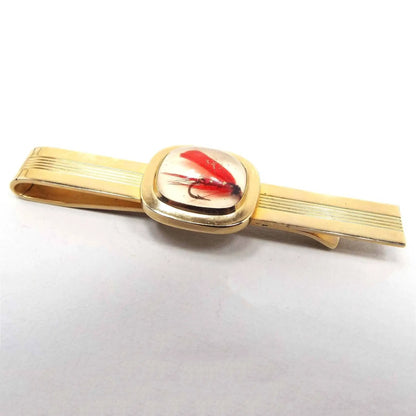 Front view of 1950's Mid Century vintage fisherman's tie bar. The metal is gold tone in color and is a wide slide on style. The middle of the front bar has a small line design texture. The middle has a rounded square with a resin cab that has a small red fly fishing lure embedded inside. The background of the cab is white and the rest of the resin is clear.
