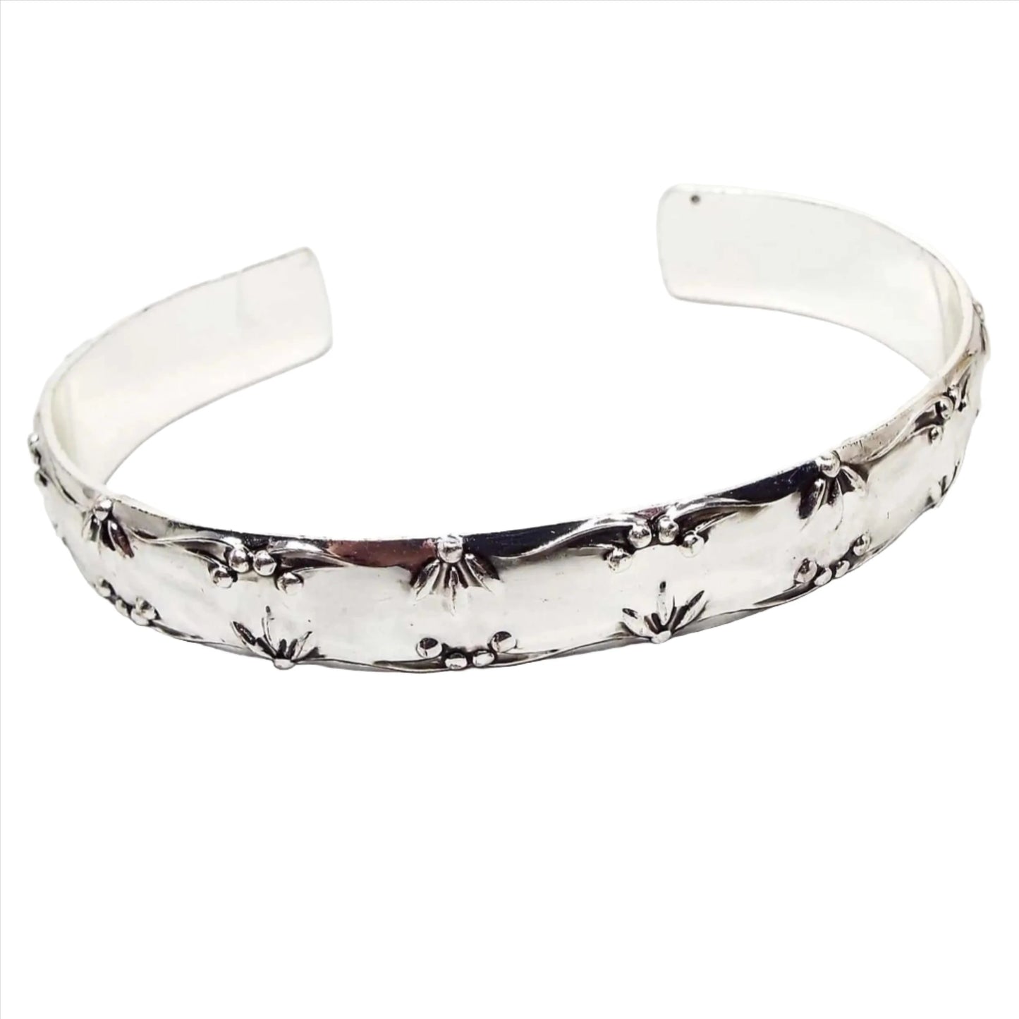 Front view of the retro 1970's Avon cuff bracelet. It is a bright shiny silver tone in color. The top and bottom edge of outside design has raised partial flower petal designs with berries in between. The inside of the cuff is flat and smooth.