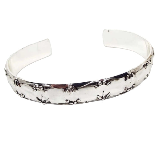 Front view of the retro 1970's Avon cuff bracelet. It is a bright shiny silver tone in color. The top and bottom edge of outside design has raised partial flower petal designs with berries in between. The inside of the cuff is flat and smooth.