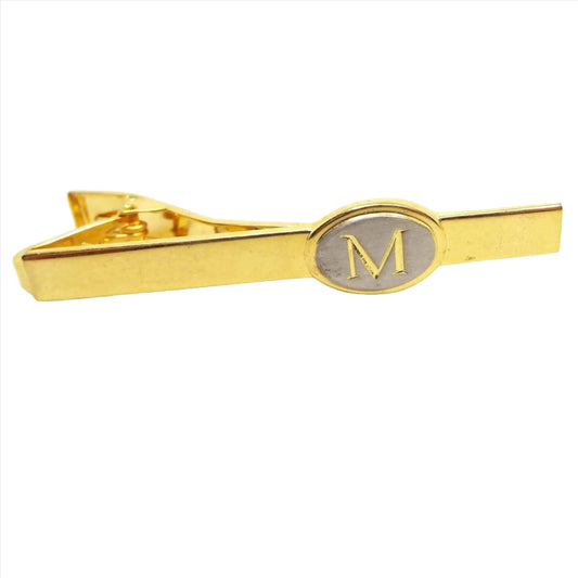 Front view of the retro vintage two tone letter tie clip bar. The main part of the tie clip is gold tone in color. Towards the end is a matte silver tone oval with a gold color block letter initial M in the middle. There is an alligator style clasp showing on the back.