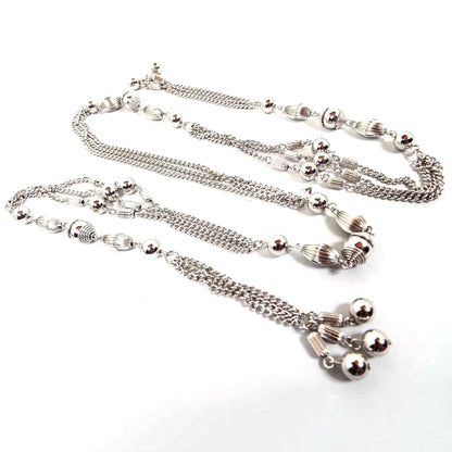 Top view of the Mid Century vintage Sarah Coventry tassel lariat necklace. It is silver tone in color with strands of curb chain in between round and corrugated metal beads. The ends have corrugated barrel beads with round smooth metal beads at the ends of the tassel areas. The necklace is open and wraps around lariat style rather than having a clasp.