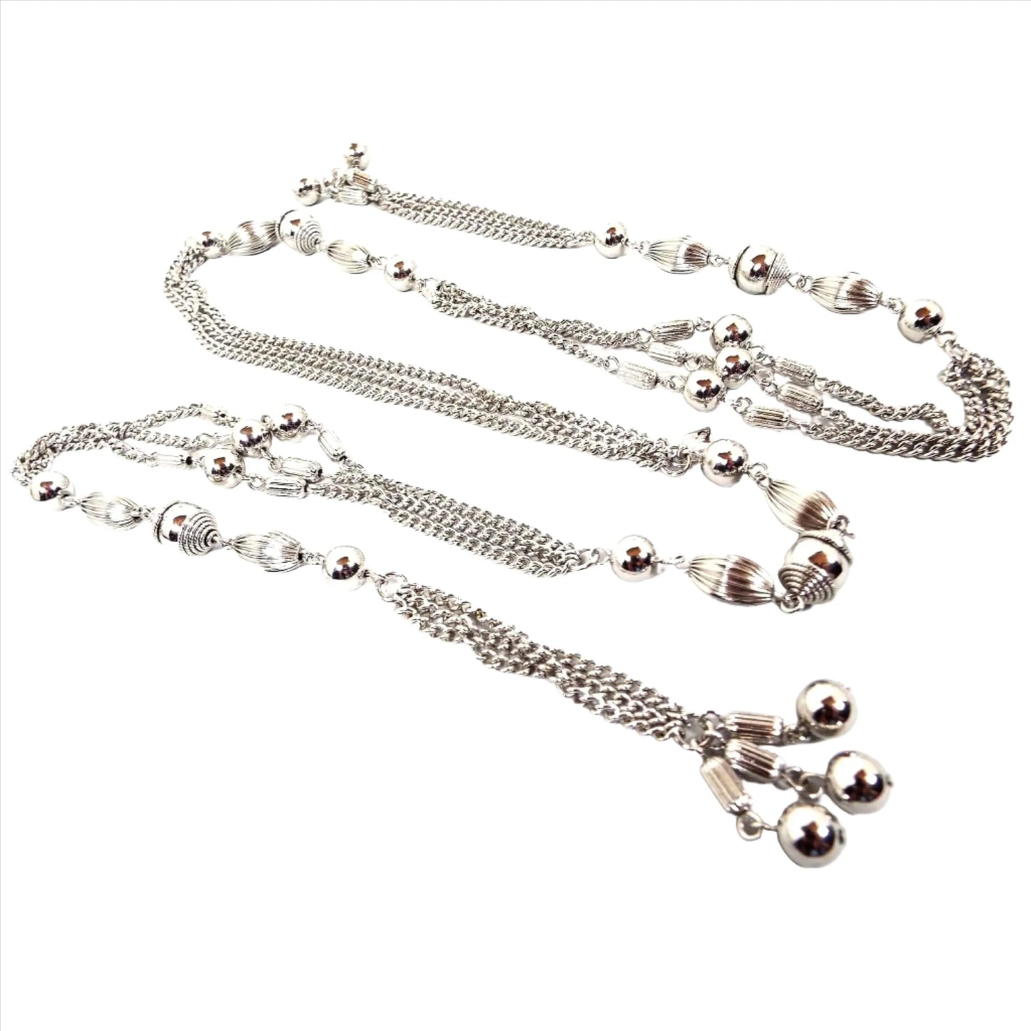 Top view of the Mid Century vintage Sarah Coventry tassel lariat necklace. It is silver tone in color with strands of curb chain in between round and corrugated metal beads. The ends have corrugated barrel beads with round smooth metal beads at the ends of the tassel areas. The necklace is open and wraps around lariat style rather than having a clasp.