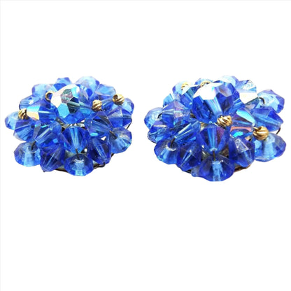 Clip on earrings are beaded in a flower like shape with faceted blue beads. Beads have AB coating on some of the facets which gives them flashes of other colors when you move around. Top beads are large ovals, followed by a row of double cone or bicone beads, and then saucer shaped beads. In between some of the beads are small gold color beads with wavy indented lines on them.