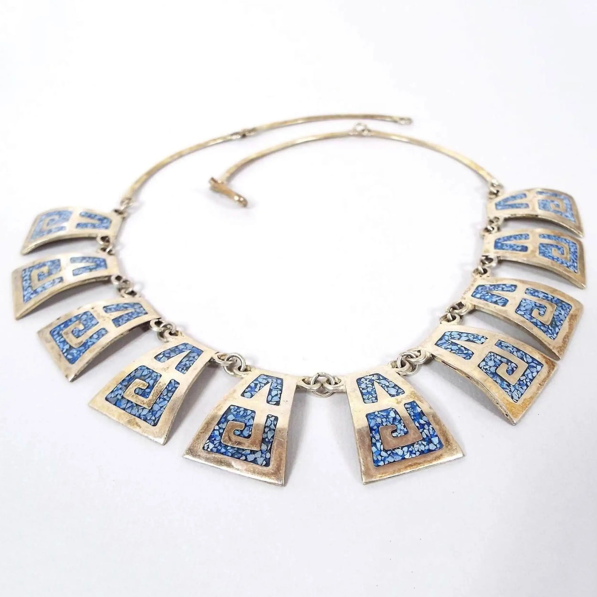 Top view of the retro vintage alpaca and sterling link necklace. The sterling links that form the top part of the necklace are curved rectangle shape with a hook clasp at the end. The bottom part has curved trapezoid shaped silver tone alpaca alloy links that are slightly curved outward. The links have a Southwestern design with tiny blue turquoise chips and resin.