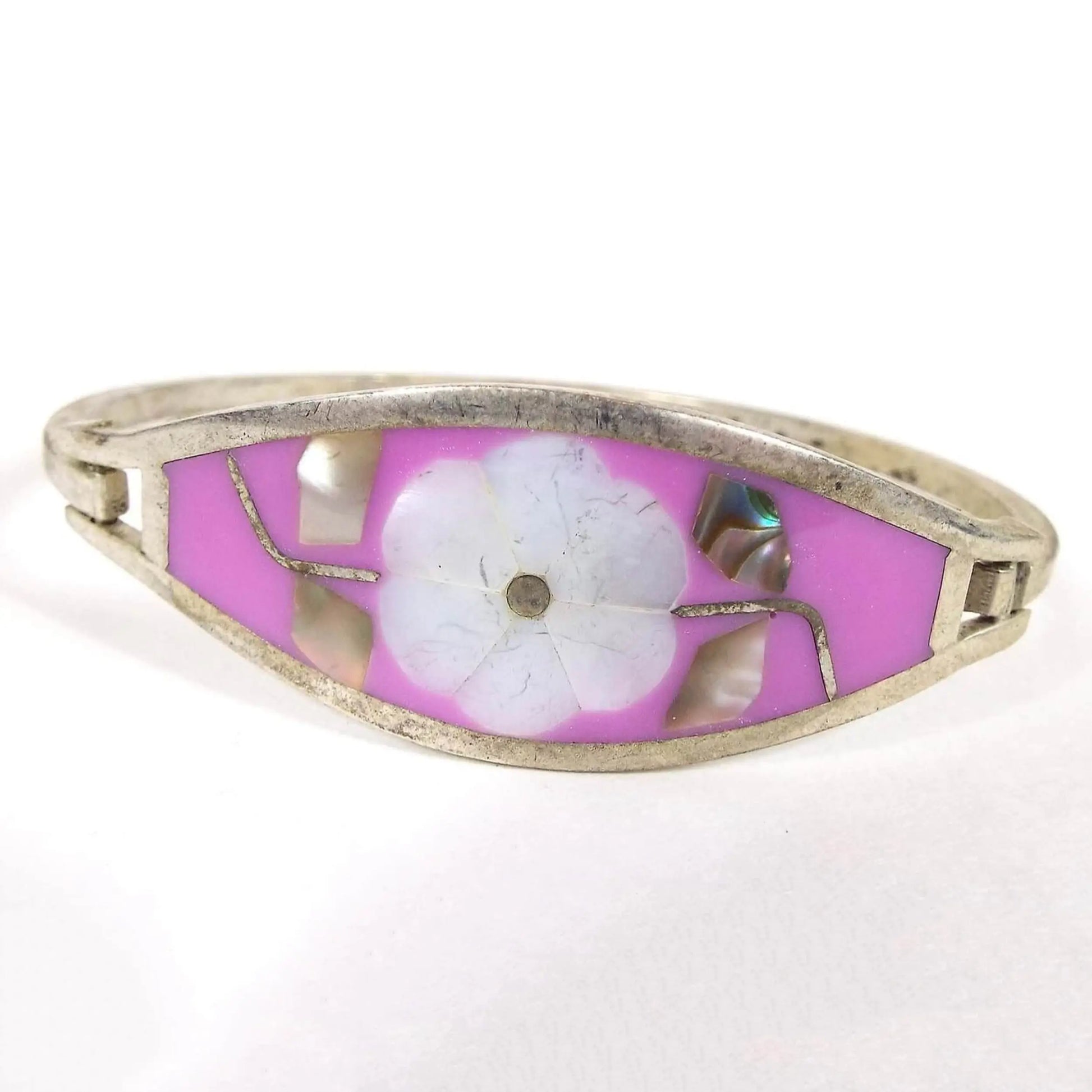 Front view of the retro vintage hinged bangle bracelet. The alpaca metal is silver tone in color. The front has a large tapered curved area with bright pink enamel and a flower design in the middle. Flower petals are pearly white inlaid mother of pearl shell and the leaves on each side of the flower are multi color pearly inlaid abalone shell. There is a thin curved band of metal forming the back of the bangle that's hinged on one side and has a hook clasp on the other to open and close the bracelet.