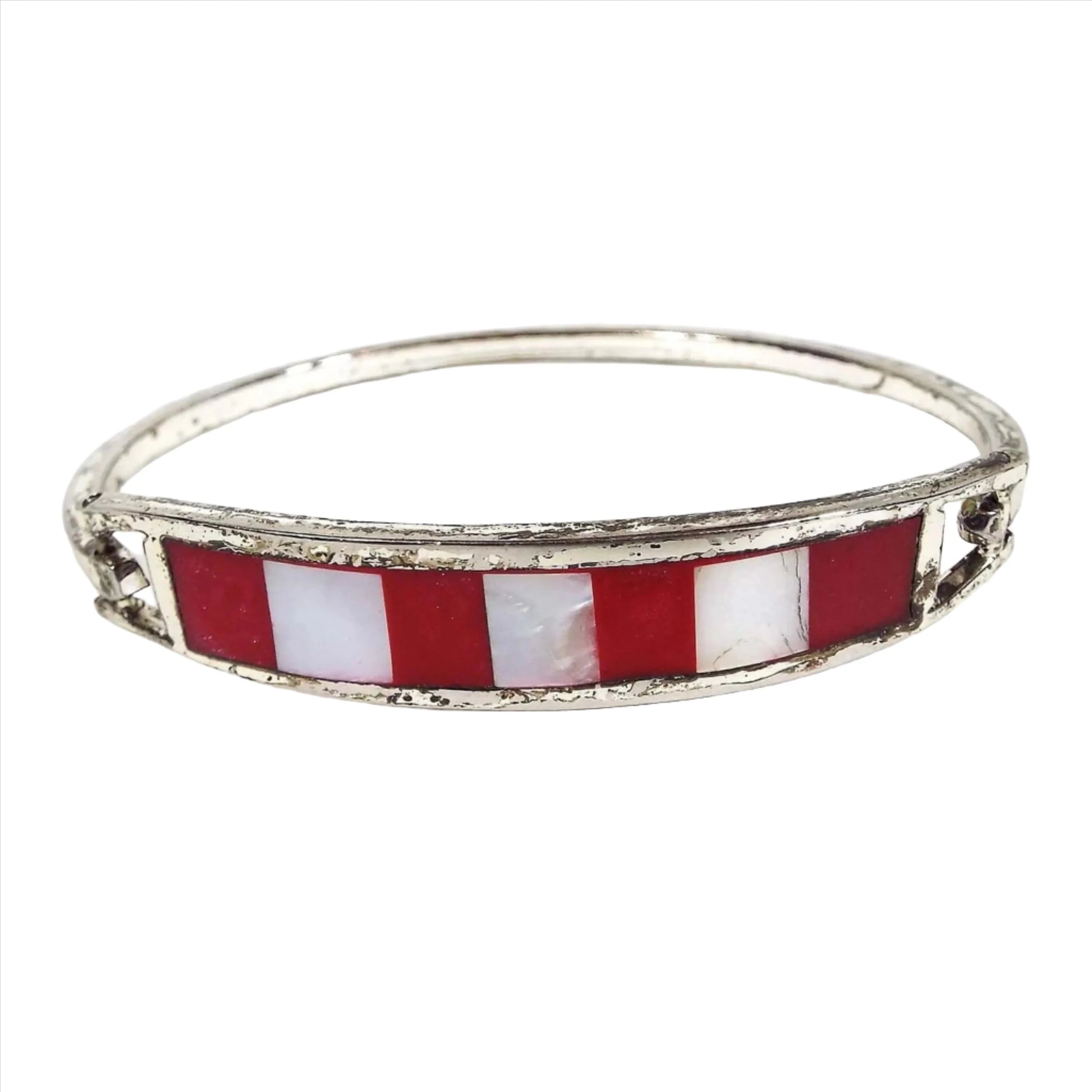 Angled front and side view of the retro vintage Mexican hinged bangle bracelet. The metal is darkened silver tone in color. The front has a curved bar with stripes of red resin enamel and pearly white inlaid mother of pearl shell. The metal band on the back is thin and squared and has a hinge on one side and a hook on the other to open and close the bangle.