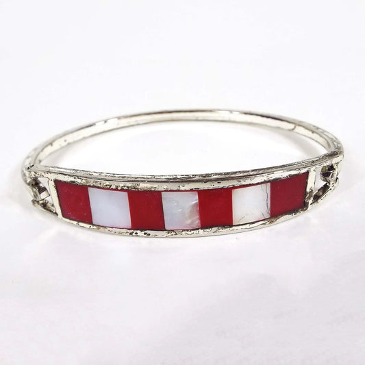 Angled front and side view of the retro vintage Mexican hinged bangle bracelet. The metal is darkened silver tone in color. The front has a curved bar with stripes of red resin enamel and pearly white inlaid mother of pearl shell. The metal band on the back is thin and squared and has a hinge on one side and a hook on the other to open and close the bangle.