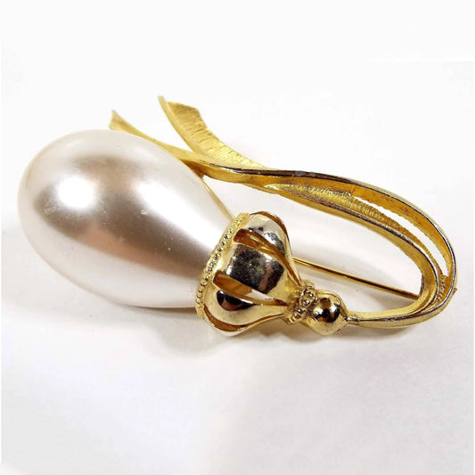 Front view of the retro vintage faux pearl brooch. The metal is gold tone in color. There is a large off white coated plastic rounded teardrop imitation pearl on the left side. Past that is a bulbous area with cut out lines down to a round ball shape and then metal shaped like two ribbons coming off the very end and curving back around to the faux pearl.