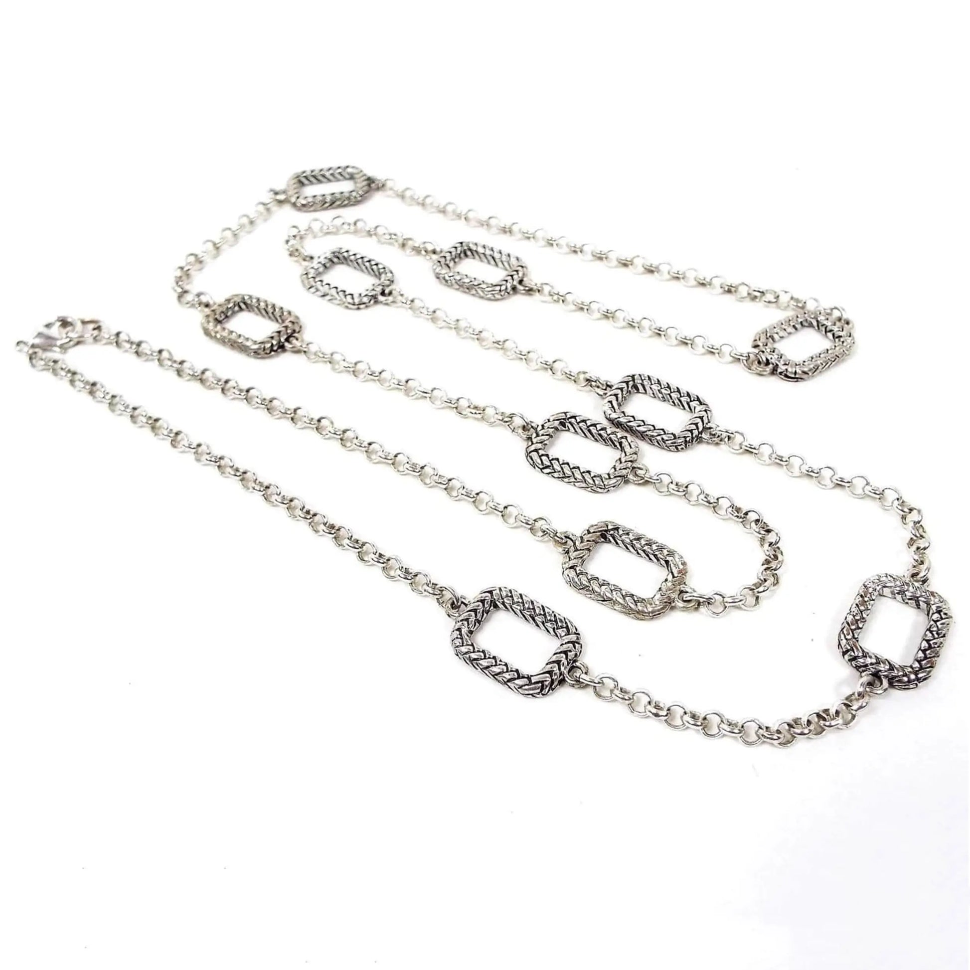 Top view of the long retro vintage chain necklace. The metal is silver tone in color. The main part of the necklace has a rolo chain link design. There are ten large rounded edge rectangles spread throughout the chain that have a textured wheat like design. There is a lobster claw clasp at the end.