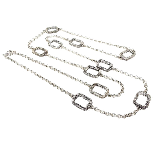 Top view of the long retro vintage chain necklace. The metal is silver tone in color. The main part of the necklace has a rolo chain link design. There are ten large rounded edge rectangles spread throughout the chain that have a textured wheat like design. There is a lobster claw clasp at the end.