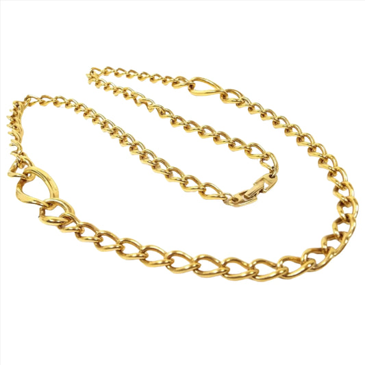 Top view of the retro vintage Napier chain necklace. It is gold tone in color with a curved oval curb chain design. There are two areas on either side that have sets of three links that are larger in size with the largest in the middle. There is a snap lock clasp on the end.