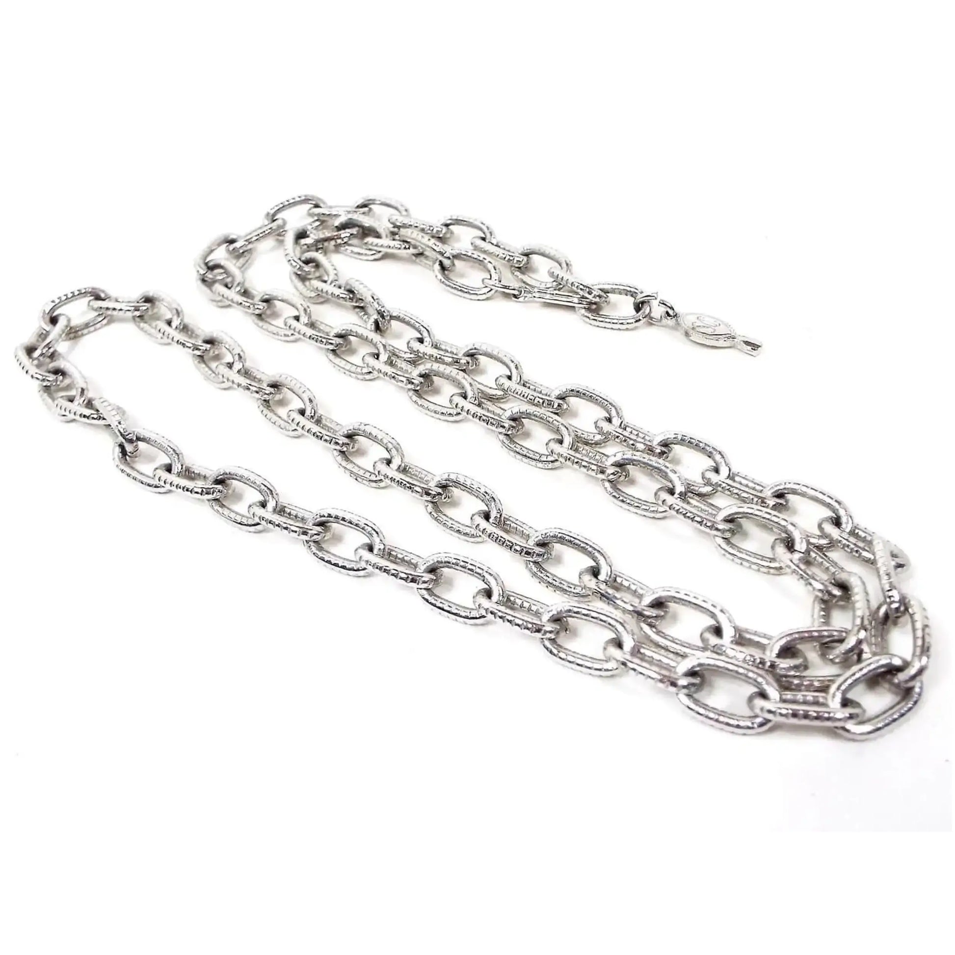 Front view of the Mid Century vintage Sarah Coventry chain necklace. It is silver tone in color. The cable chain has larger sized oval links with a lightly textured tiny line design on them. There is a hook style clasp at one end and an open ring at the other so you can adjust the length when wearing. There is a hang tag at one end with SC stamped on it.
