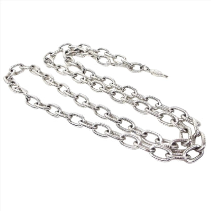 Front view of the Mid Century vintage Sarah Coventry chain necklace. It is silver tone in color. The cable chain has larger sized oval links with a lightly textured tiny line design on them. There is a hook style clasp at one end and an open ring at the other so you can adjust the length when wearing. There is a hang tag at one end with SC stamped on it.