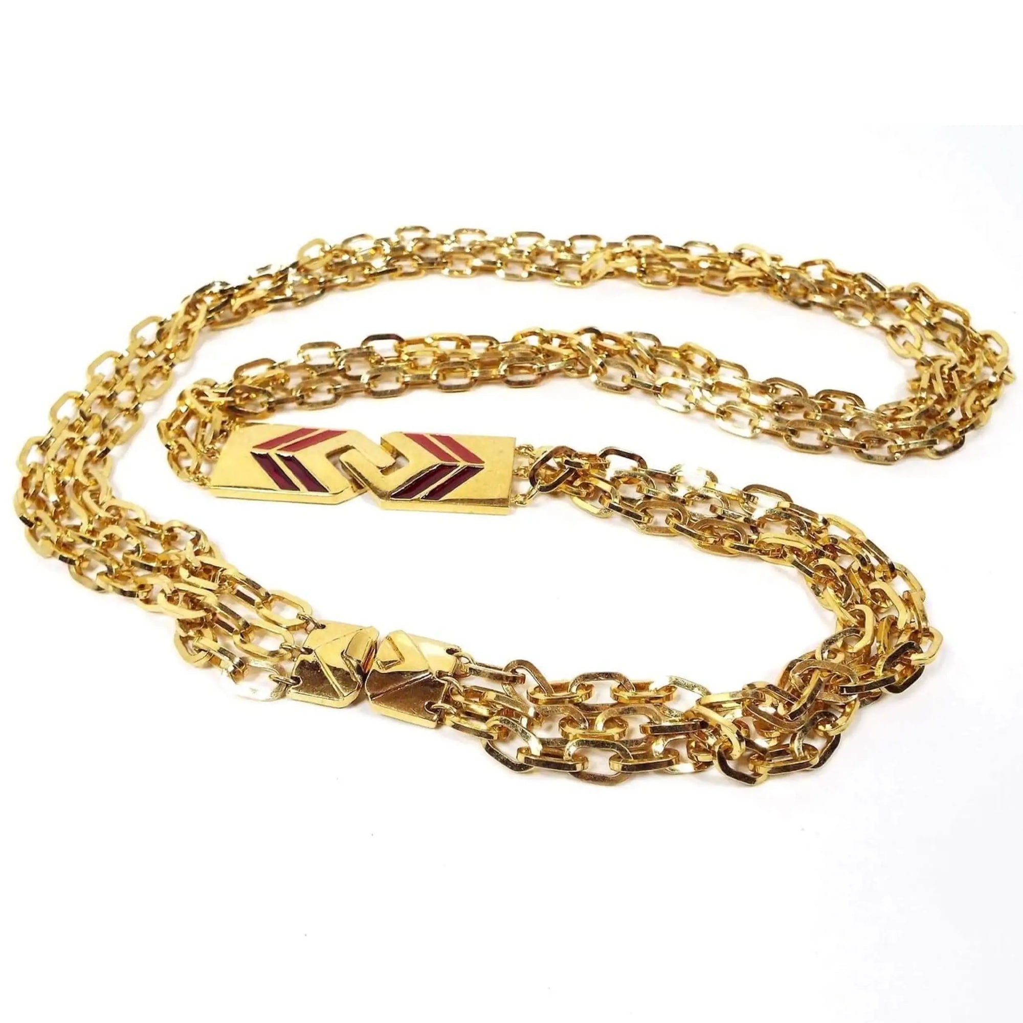 Top view of the retro vintage Monet chain necklace. The plated metal is gold tone in color. The necklace is long with 3 strands of oval cable link chain. There is a larger box clasp at the end of the necklace. Part way down the necklace are two large interlocking links that form a long rectangle. Each link has a chevron double V style design. One side of the chevron has red enamel and the other has dark red enamel.