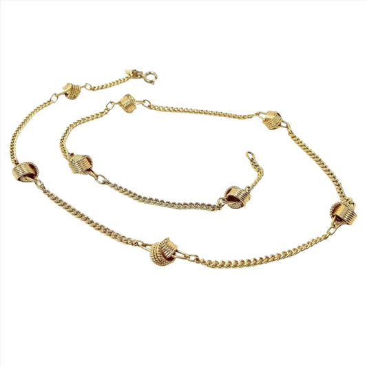 Top view of the retro vintage Avon knot chain necklace. It has a thinner style curb link chain with a round spring ring clasp at the end. There are spaced round textured open rings that are joined together to form the knot areas spaced out on the chain. There are 8 knot areas. The entire necklace is gold tone in color. 