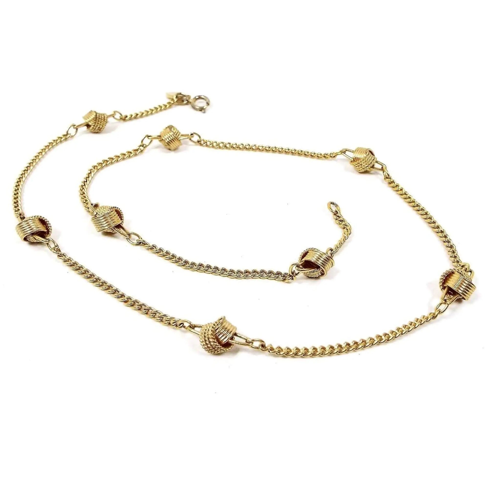 Top view of the retro vintage Avon knot chain necklace. It has a thinner style curb link chain with a round spring ring clasp at the end. There are spaced round textured open rings that are joined together to form the knot areas spaced out on the chain. There are 8 knot areas. The entire necklace is gold tone in color. 
