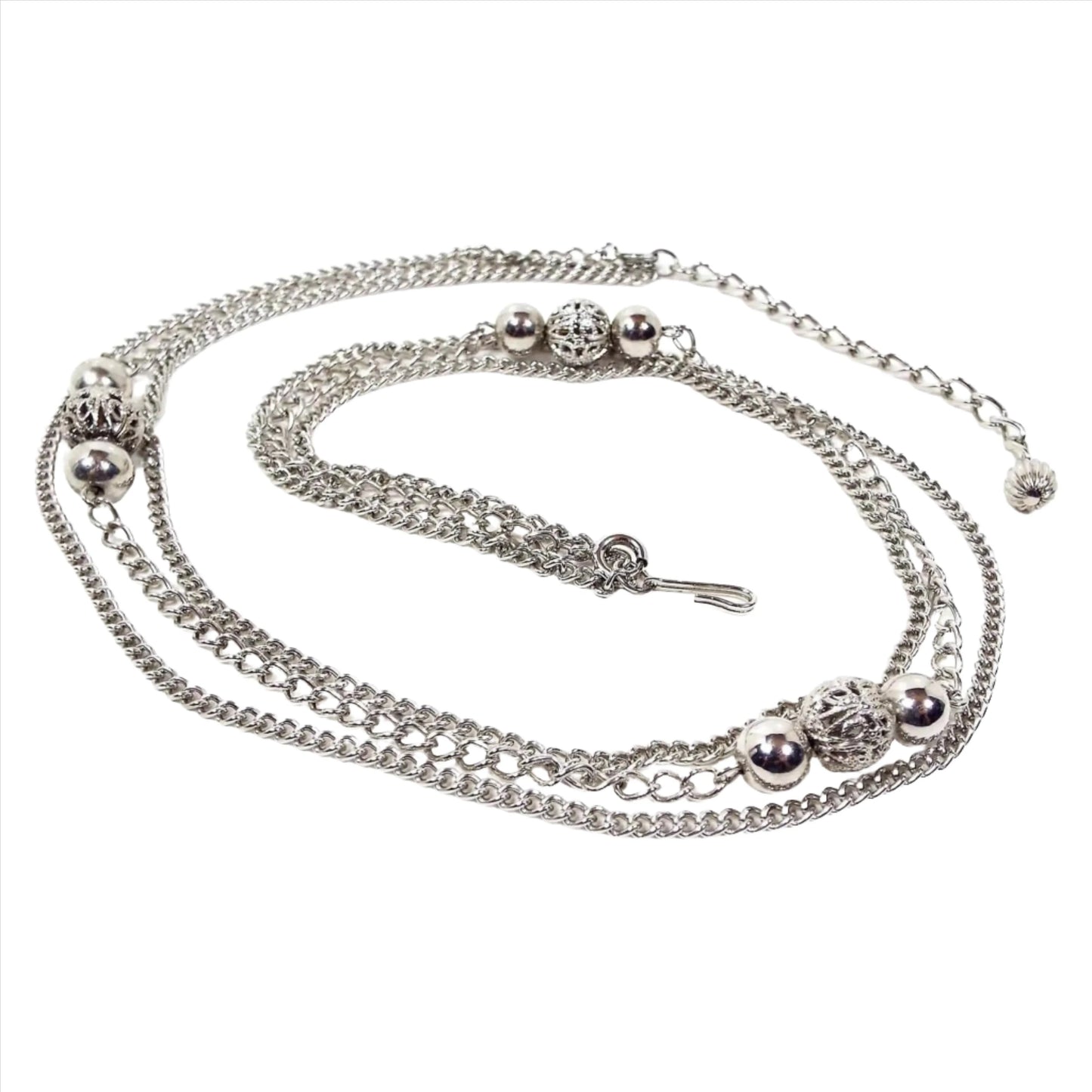 Top view of the retro vintage multi strand silver tone chain necklace. The top and bottom strands have small curb link chain. The middle strand has a wider curb link chain and three sets of metal bead clusters that have shiny round beads with filigree beads in the middle. It has spring ring clasps on the ends of the middle strand so you can remove it if desired. There is a hook clasp at the end. The other end is wider curb links with a corrugated bead at the end so you can adjust length when worn.