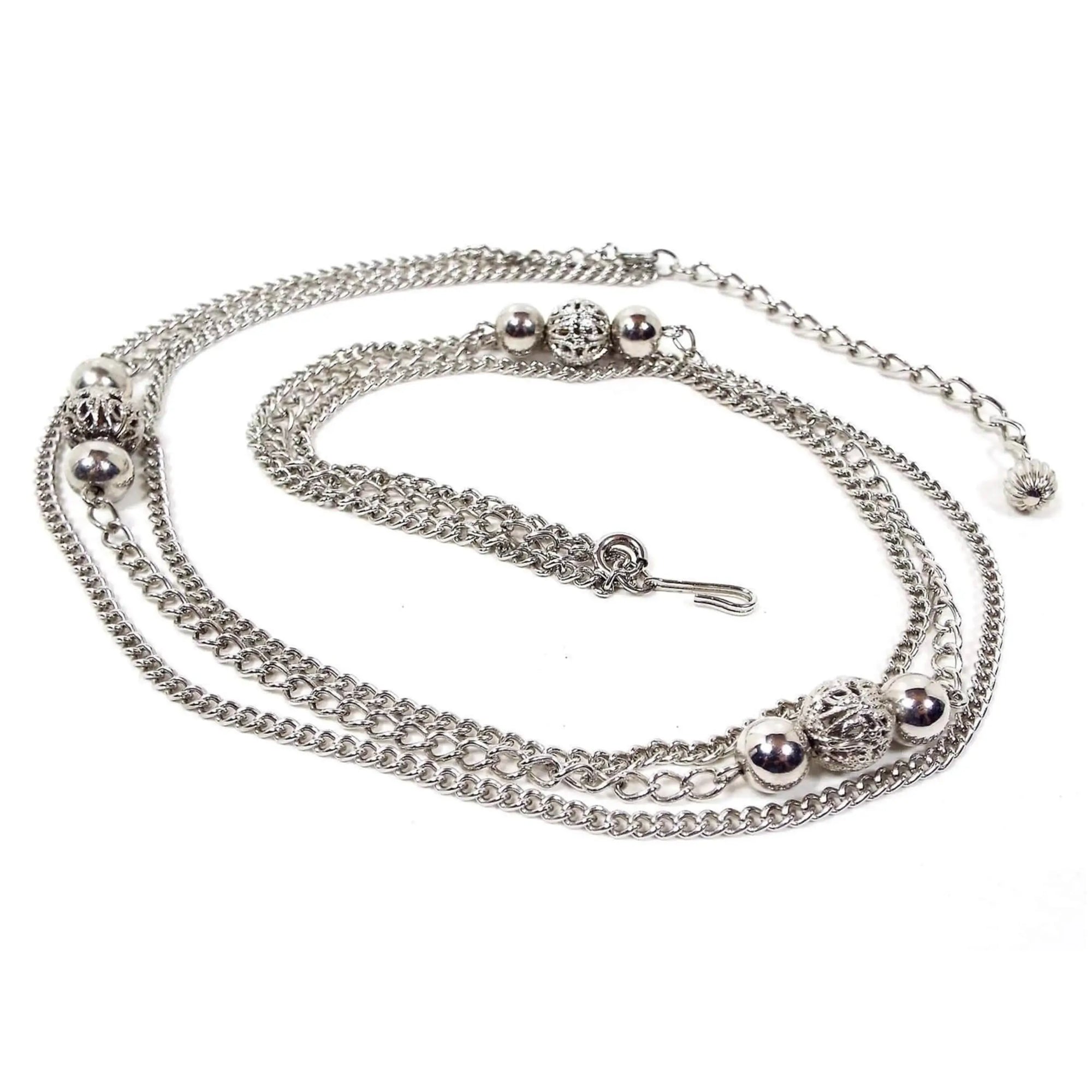 Top view of the retro vintage multi strand silver tone chain necklace. The top and bottom strands have small curb link chain. The middle strand has a wider curb link chain and three sets of metal bead clusters that have shiny round beads with filigree beads in the middle. It has spring ring clasps on the ends of the middle strand so you can remove it if desired. There is a hook clasp at the end. The other end is wider curb links with a corrugated bead at the end so you can adjust length when worn.