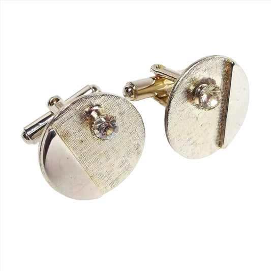 Angled front and side view of the retro vintage rhinestone cufflinks. They are round in shape. The fronts have a textured brushed matte light gold tone color with a small round bezel set rhinestone at the top. There is a raised half moon shape on one side that has silver tone color on the front. The rounded ends of the levers on the back can be seen in the photo.