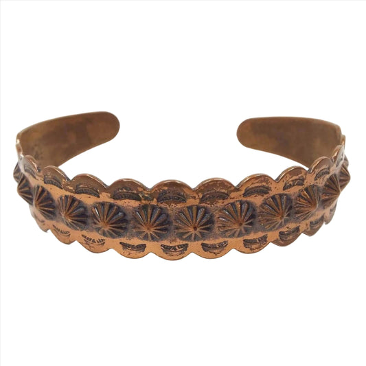 Front view of the retro vintage Bell Trading Post cuff bracelet. The bracelet is made of copper and has a repousse design where the metal has been stamped so that the front is raised up and the back of the cuff is indented in with the pattern. The top and bottom edge are scalloped with a sliver moon design and the middle of the cuff has a round raised starburst or flower like design. The ends of the cuff are tapered and rounded.