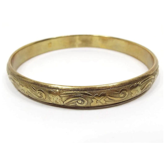 Angled side and top view of the retro vintage floral bangle bracelet. It's made of brass and is a darker gold tone in color. The bangle is round with a raised pattern of flowers against a background of lines.