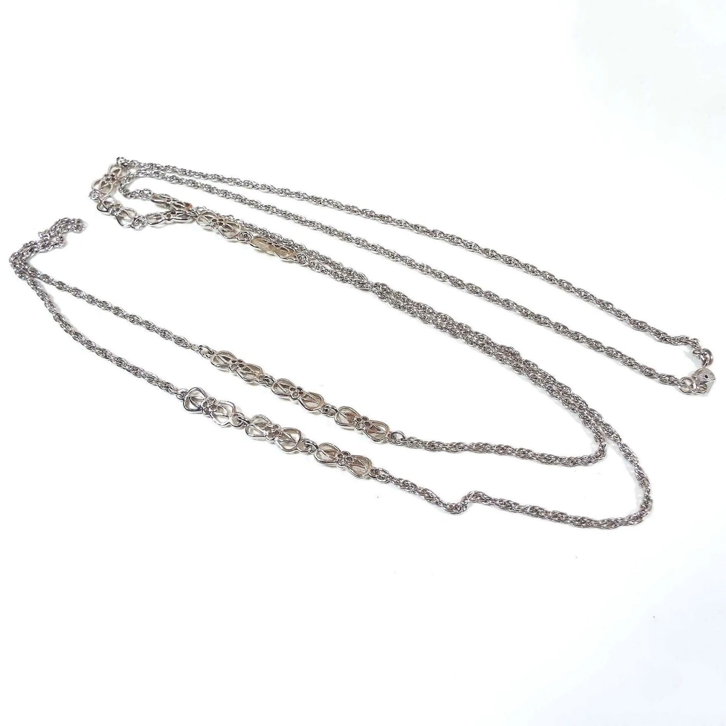 Top view of the retro vintage Monet chain necklace. The metal is silver tone in color. It has a long rope chain with two sets of spaced apart fancy links on each side for a total of 4 sets of fancy links. The fancy links are shaped like an open bow tie with an elongated open diamond shape inside that and then finally a small flower in the middle with four open petals. There is a hinged clip clasp on the end.