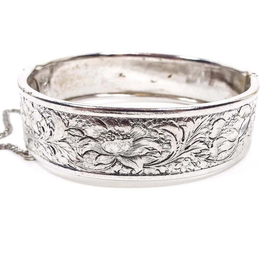 Side view of the Mid Century vintage Coro Pegasus hinged bangle bracelet. It is silver tone in color and has an embossed floral design all the way around the bracelet. It is wider style with a small safety chain by the clasp. The inside of the bangle has some darkened spots from age.