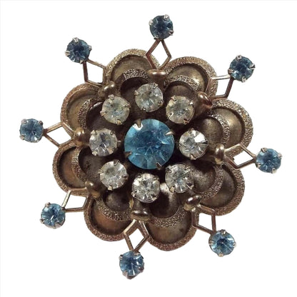 Front view of the 1950's Mid Century vintage rhinestone brooch pin. Brooch has a flower like starburst design. Middle rhinestone is round with light blue color and is surrounded by smaller round clear rhinestones. Some of the clear rhinestones are lightly cloudy and not perfectly clear. There are two layers of small rounded petals around the edge with textured edges. The bottom layer of petals has an open diamond shape in between each petal with a small round light blue rhinestone at the end of each.