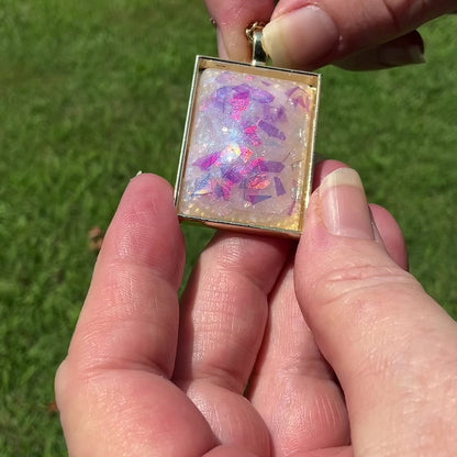 Video showing the flash and sparkle of the handmade purple, pink, and blue glitter resin pendant necklace. It is rectangle in shape with gold tone plated setting.  A hand is holding it and moving it around in the light.