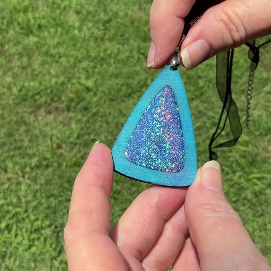 Handmade Blue Dyed Triangle Wood Pendant with Iridescent Glitter Resin video showing how the holographic glitter in the middle sparkles and changes colors as it moves around in the light.