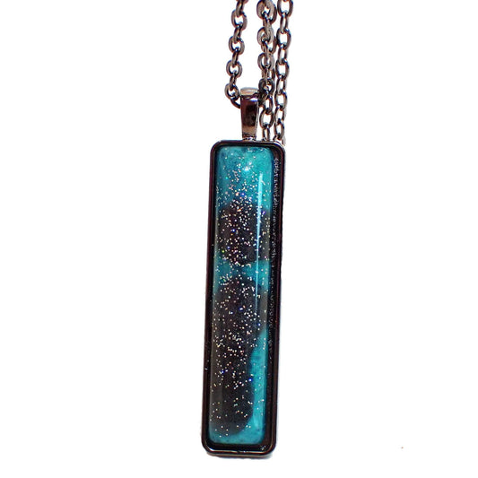 Enlarged front view of the handmade resin bar pendant necklace. The metal is gunmetal gray in color. There is a long rectangle bar pendant with rounded corners. The front resin cab has marbled shades of pearly dark gray and teal blue. There is silver holographic glitter embedded in the resin for bits of sparkle and flashes of color as you move around in the light.