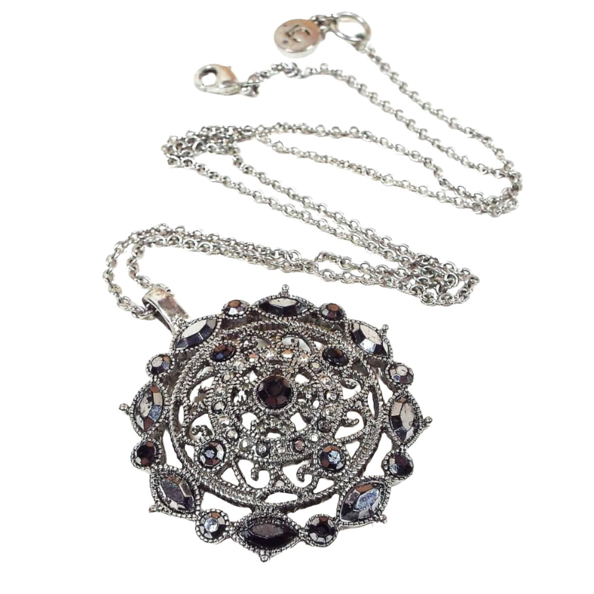 Front view of the retro vintage Liz Claiborne pendant necklace. The chain is silver tone in color. The pendant is antiqued silver tone in color with a round filigree design and various sizes and shapes of metallic gray faux marcasite rhinestones. There is a lobster claw clasp at the end and a hang tag with LC on it.