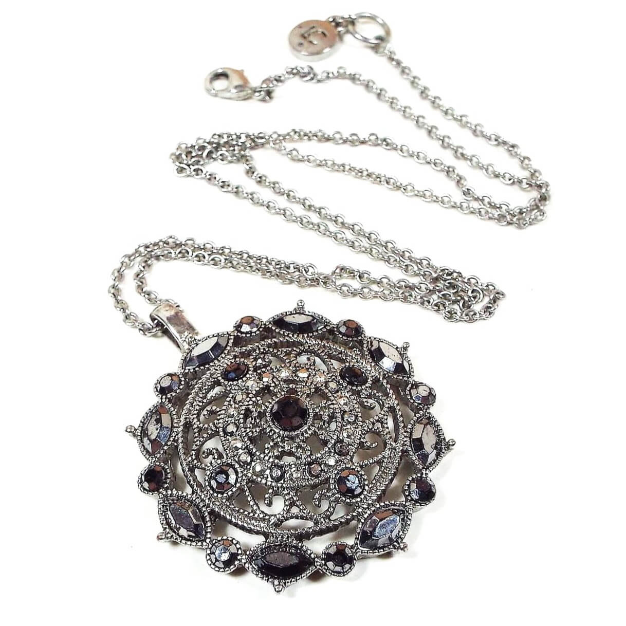 Front view of the retro vintage Liz Claiborne pendant necklace. The chain is silver tone in color. The pendant is antiqued silver tone in color with a round filigree design and various sizes and shapes of metallic gray faux marcasite rhinestones. There is a lobster claw clasp at the end and a hang tag with LC on it.