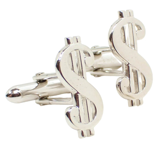Enlarged angled front view of the Mid Century vintage Swank cufflinks. They are silver tone in color and are shaped like dollar signs. 