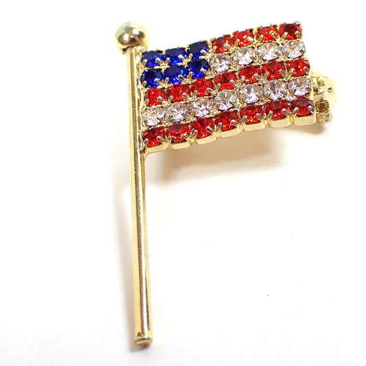 Enlarged front view of the retro vintage rhinestone flag pin. The metal is gold tone plated in color. The rhinestones are red white and blue like a US flag. 