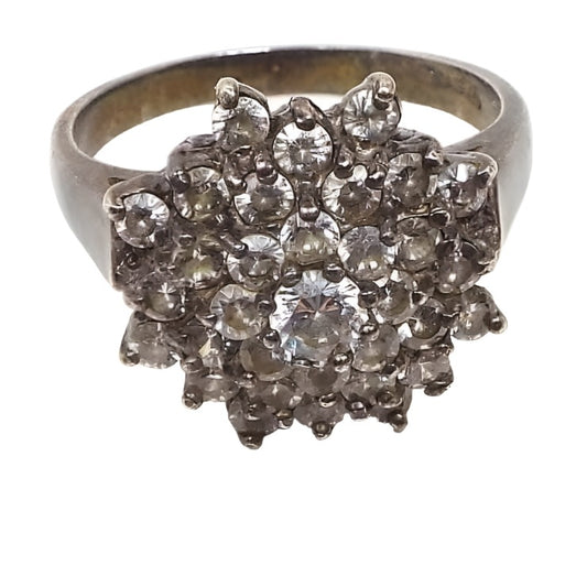 Front view of the retro vintage sterling silver cubic zirconia cocktail ring. The tope has a large flower like cluster of small round prong set CZ stones. There is a larger one in the very middle. The sterling is slightly darkened from age.